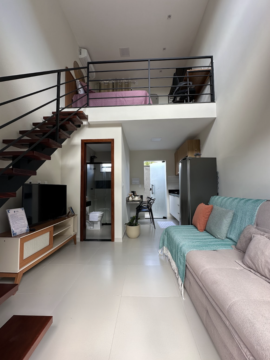 Sala do Loft Duplex com sofá bege, manta teal, escada de madeira e mezanino com cama rosê visível ao fundo