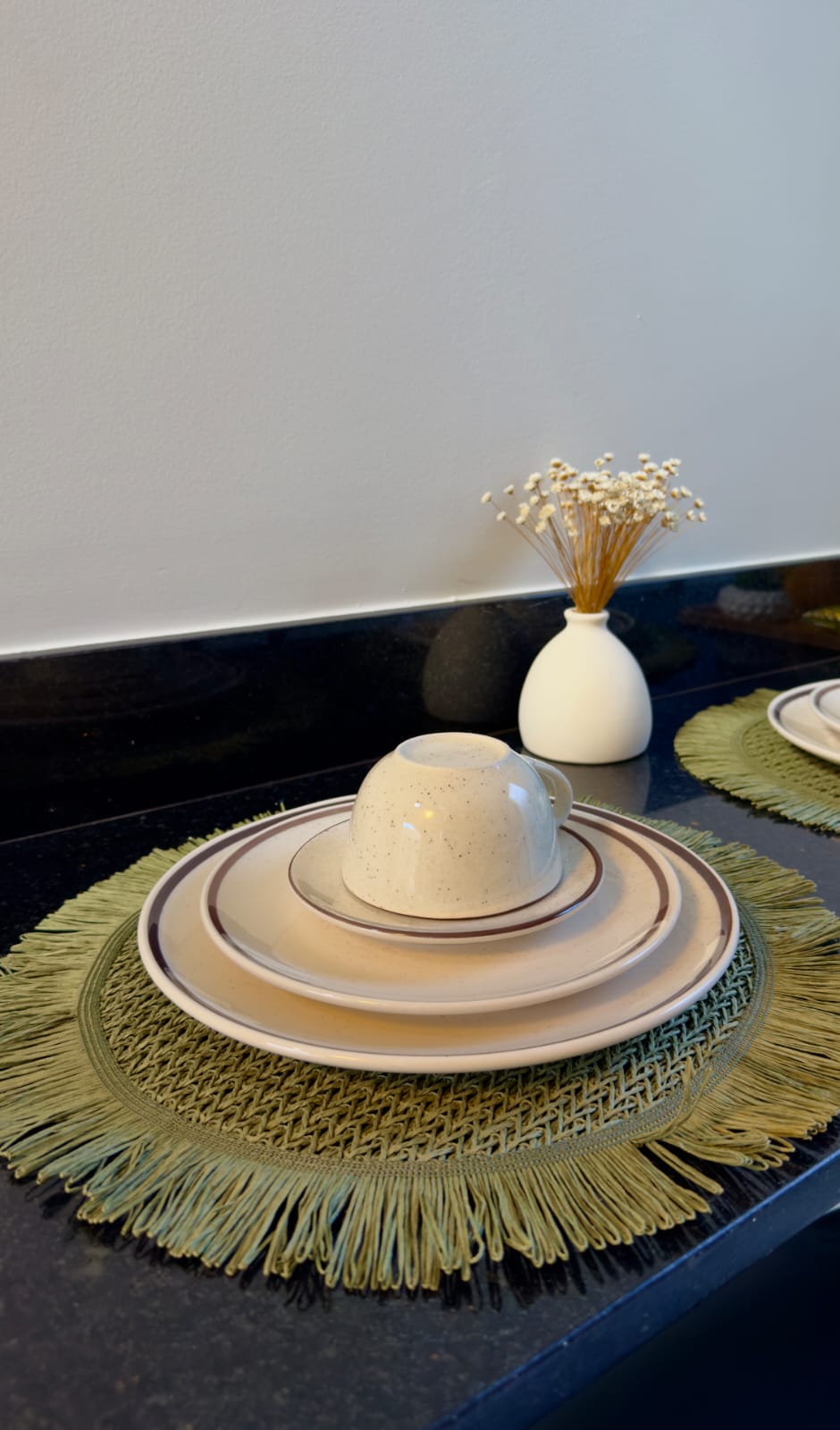 Mesa posta com jogo americano de fibra verde franjado e mini vaso com flores secas no granito