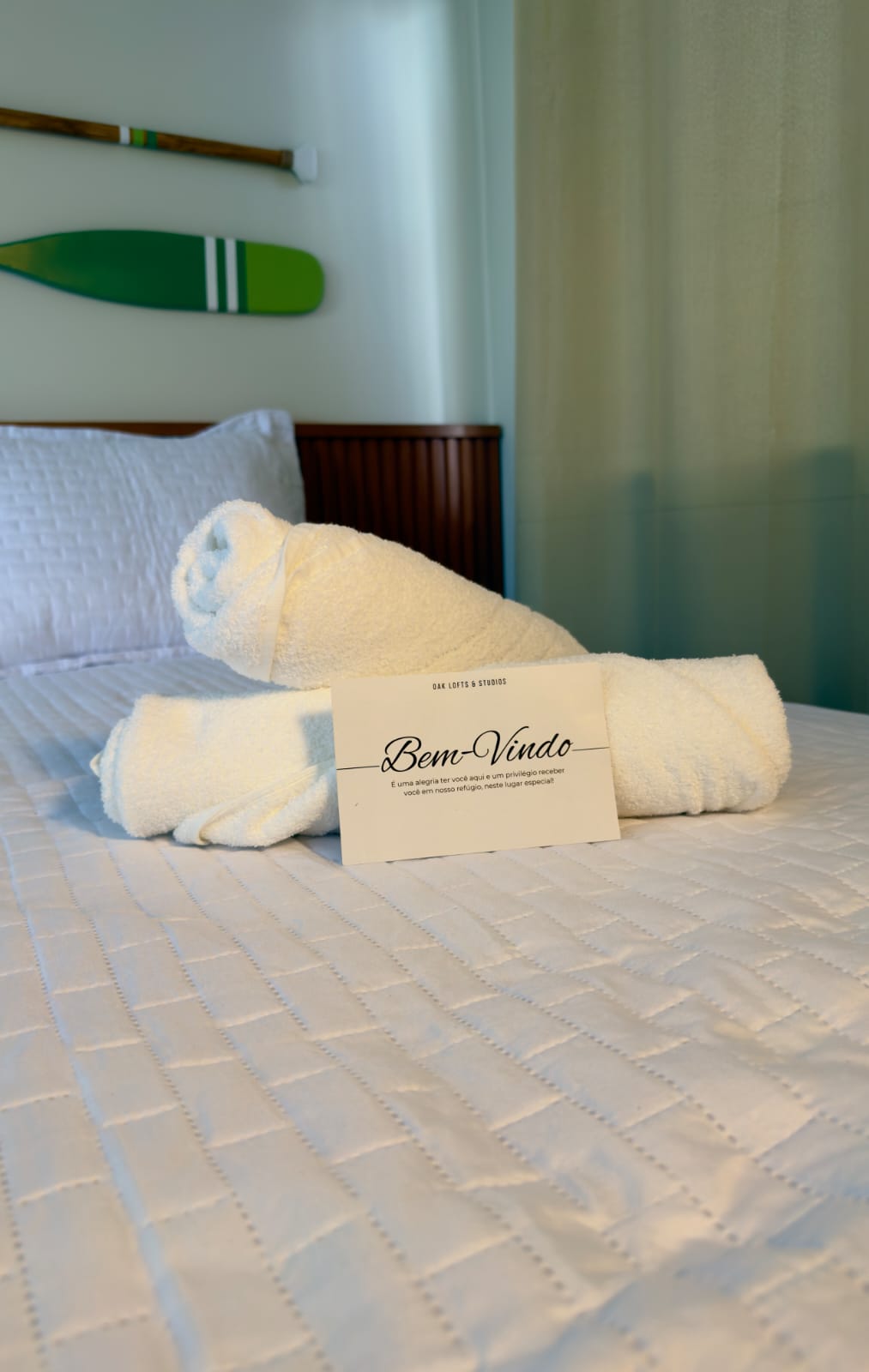 Cama branca com toalha dobrada e cartão Bem-Vindo, remos decorativos verdes na parede