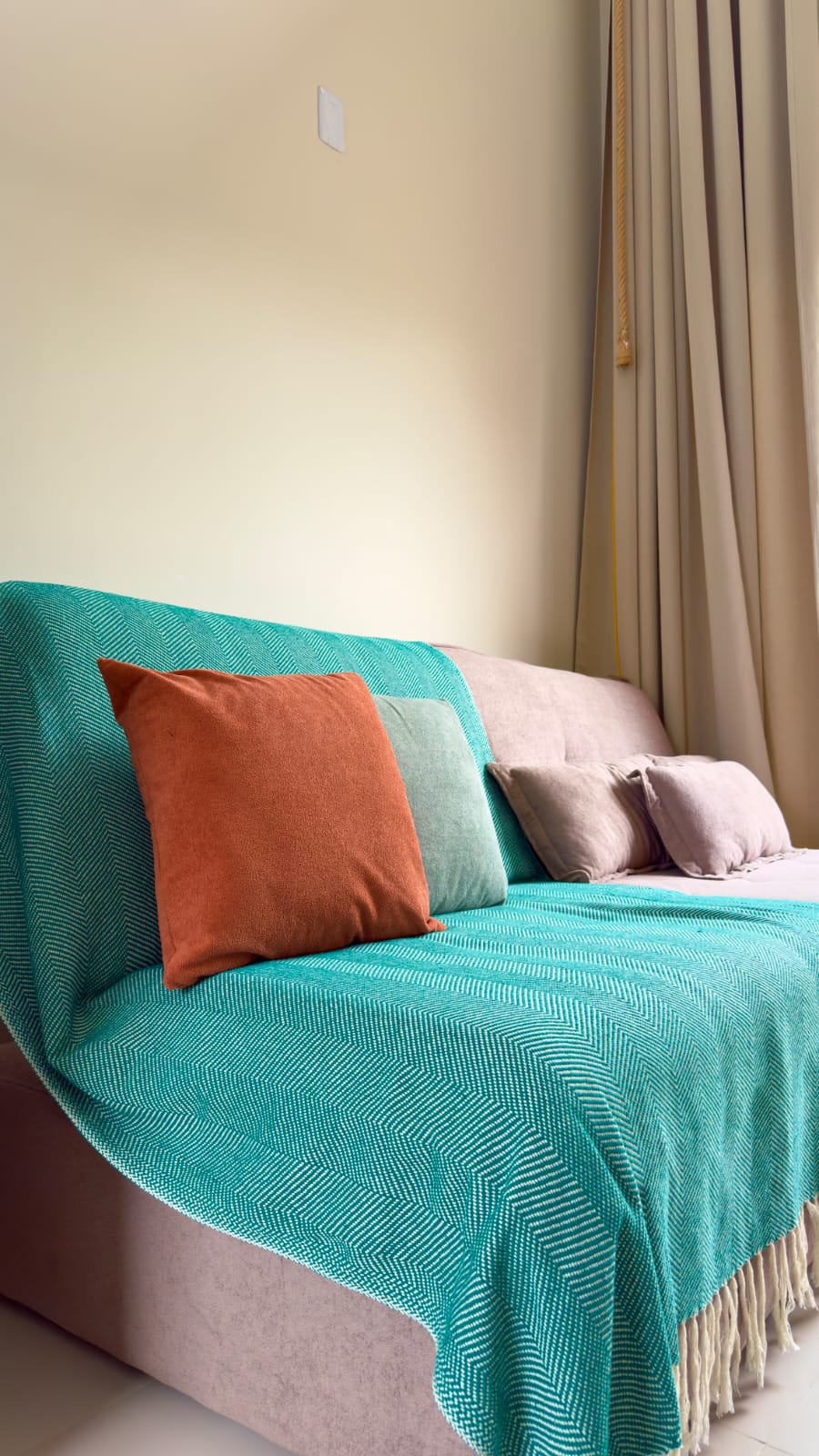 Sofá-cama com manta teal e almofada laranja com cortina bege ao fundo
